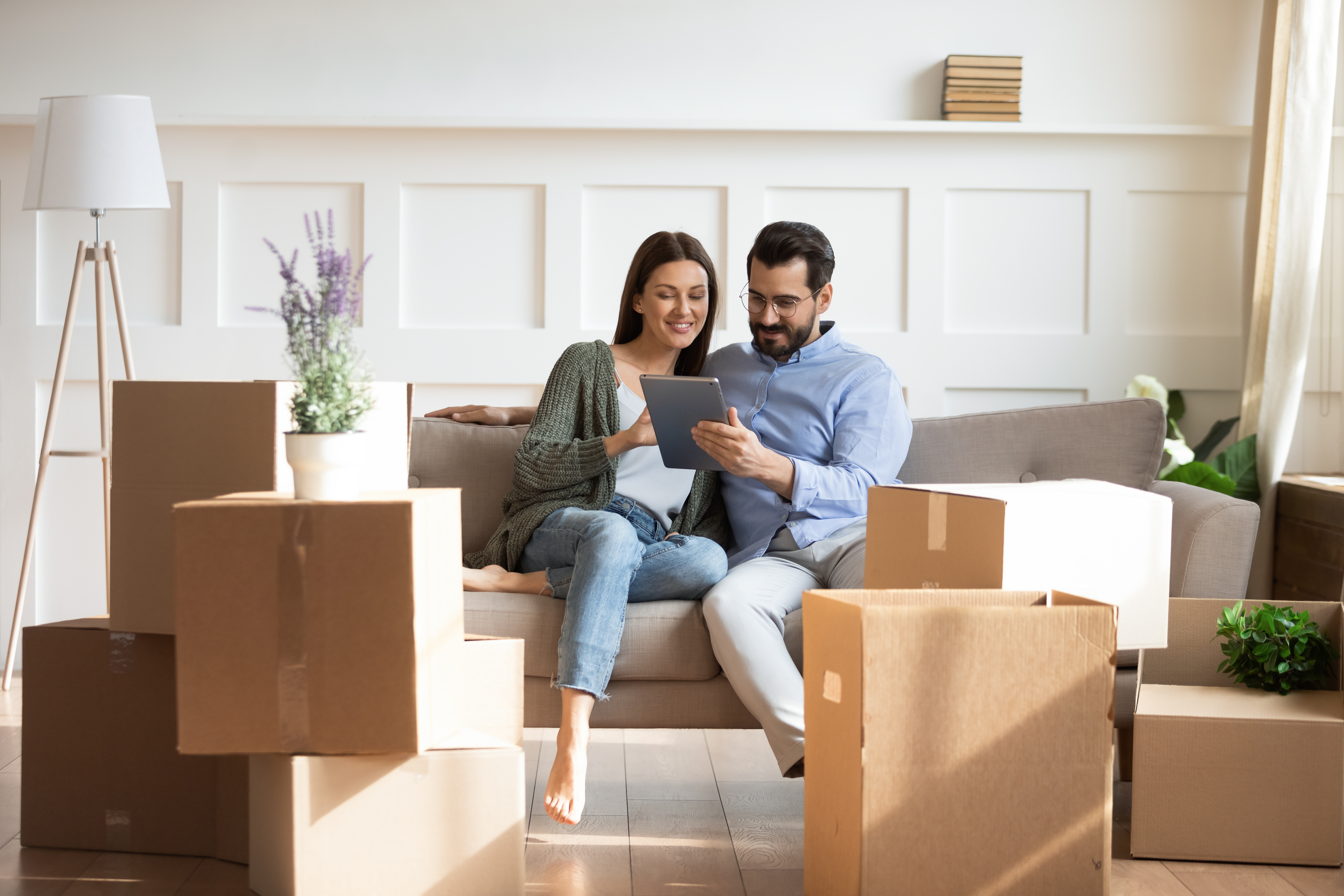 Man and woman sitting on a couch surrounded by cardboard boxes looking at a tablet