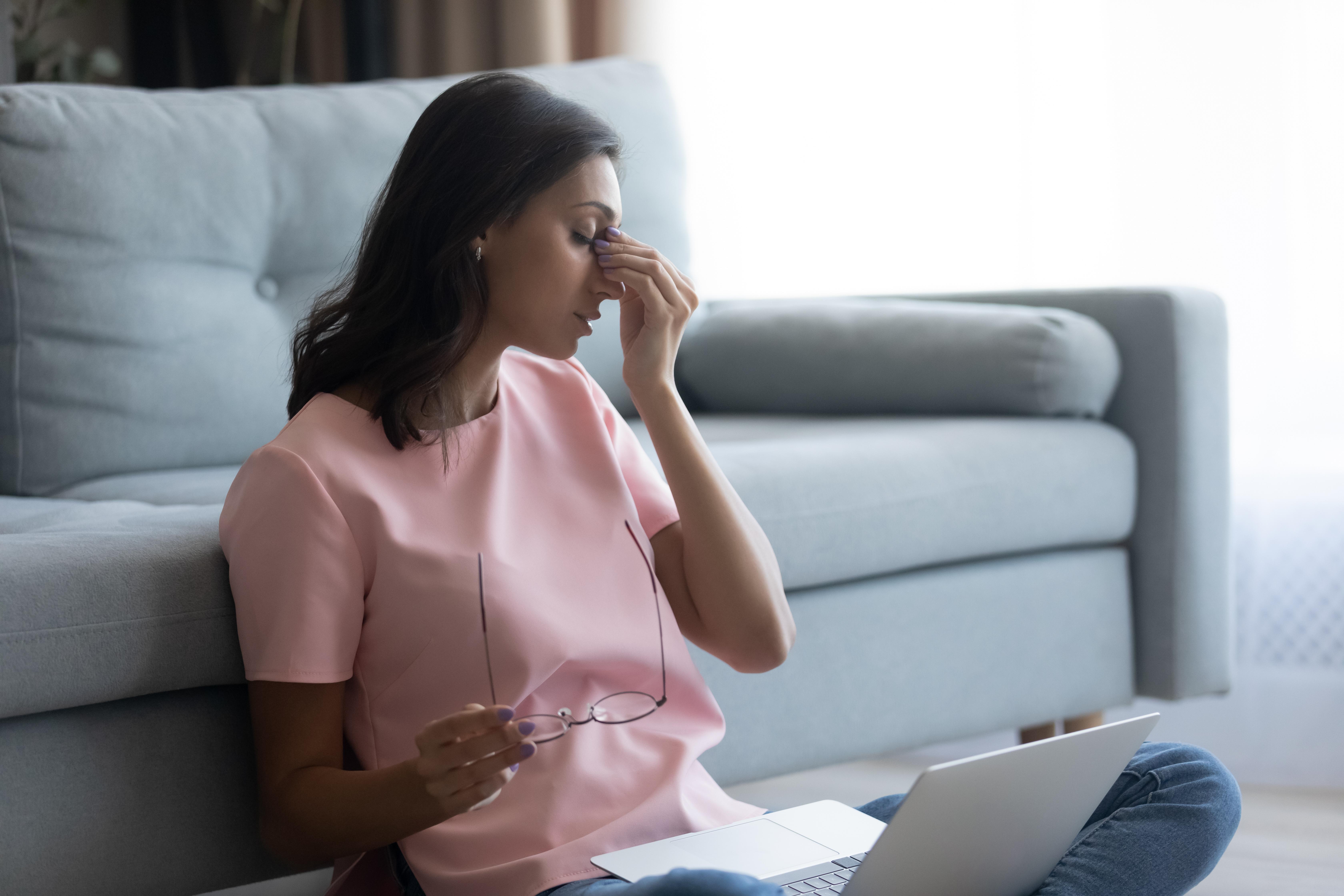 Woman sitting with a computer in her lap holding glasses and pinching the bridge of her nose