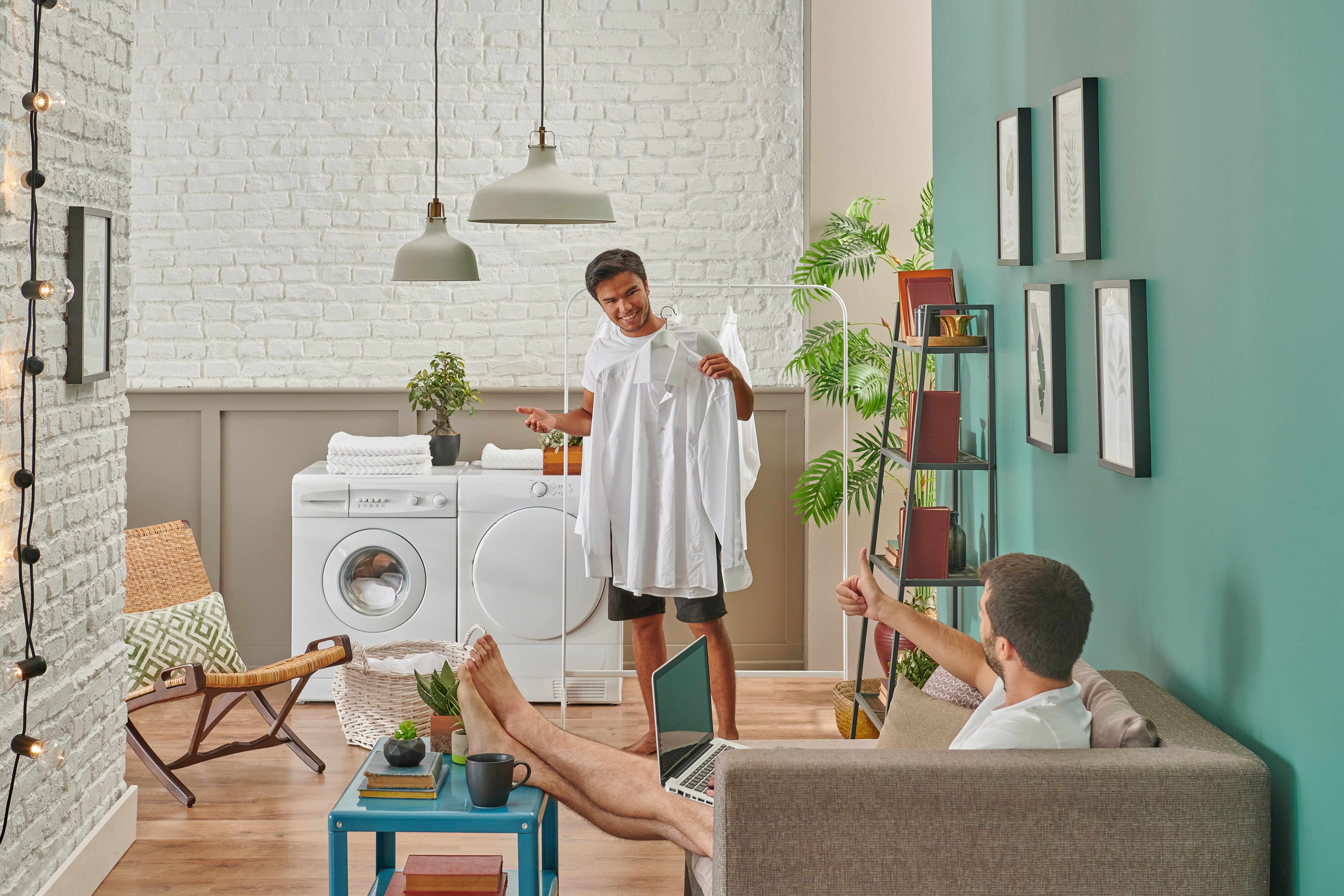 Man holding up a white button down shirt on a hanger and man sitting on the couch giving a thumbs up