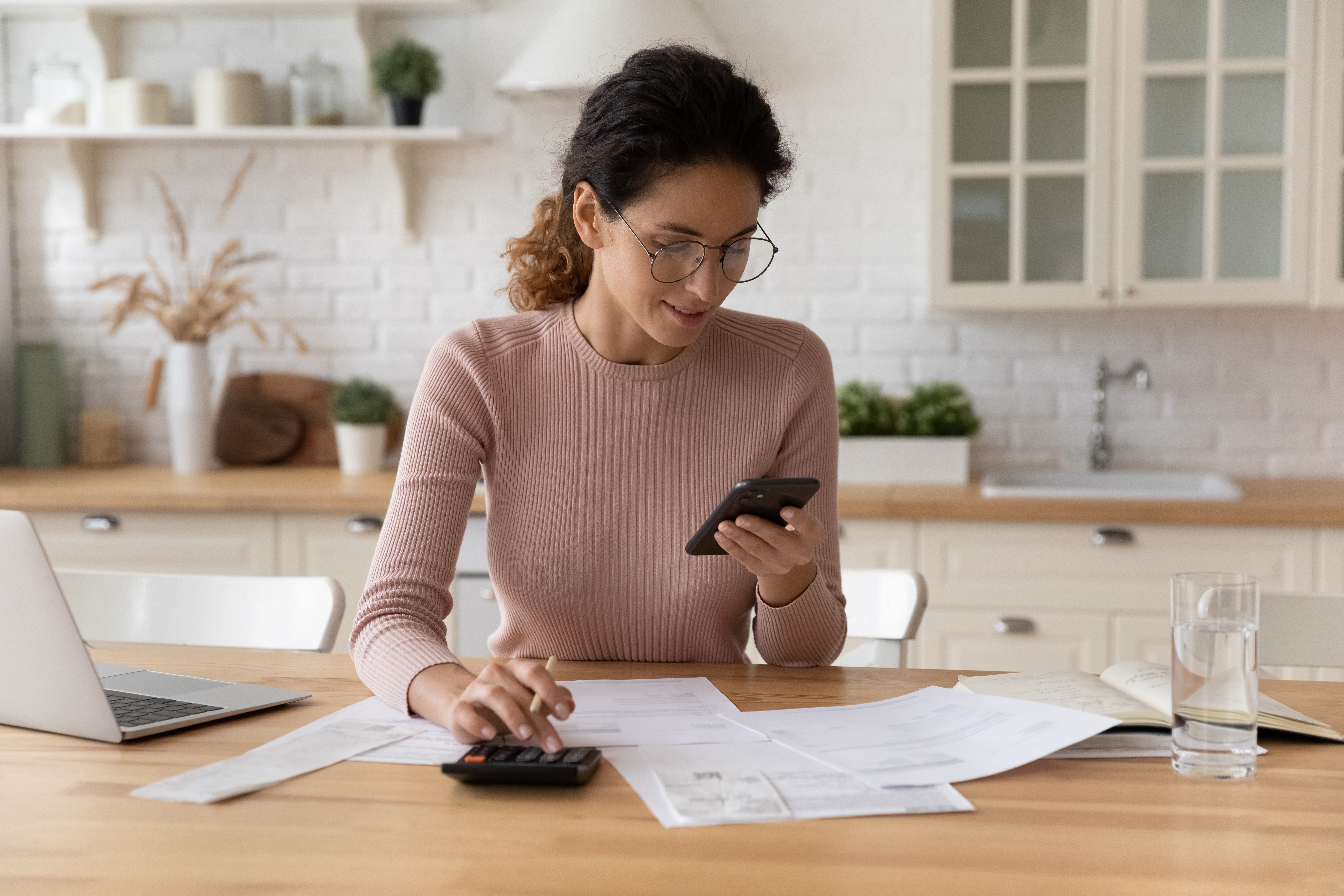 Woman sitting at a table looking at her smartphone and typing on a calculator