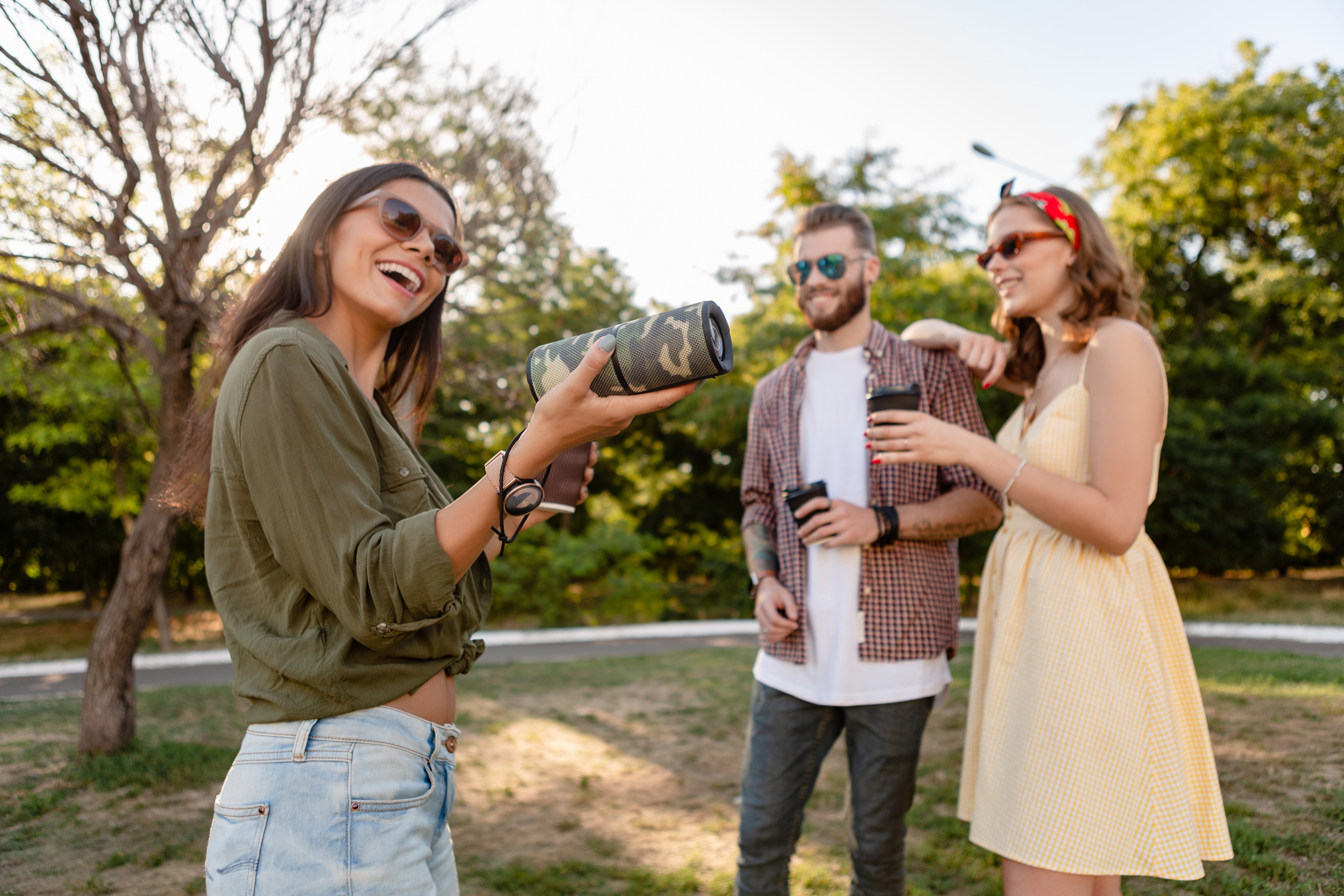 Two women and a man listening to a camouflage-colored portable speaker in a park 