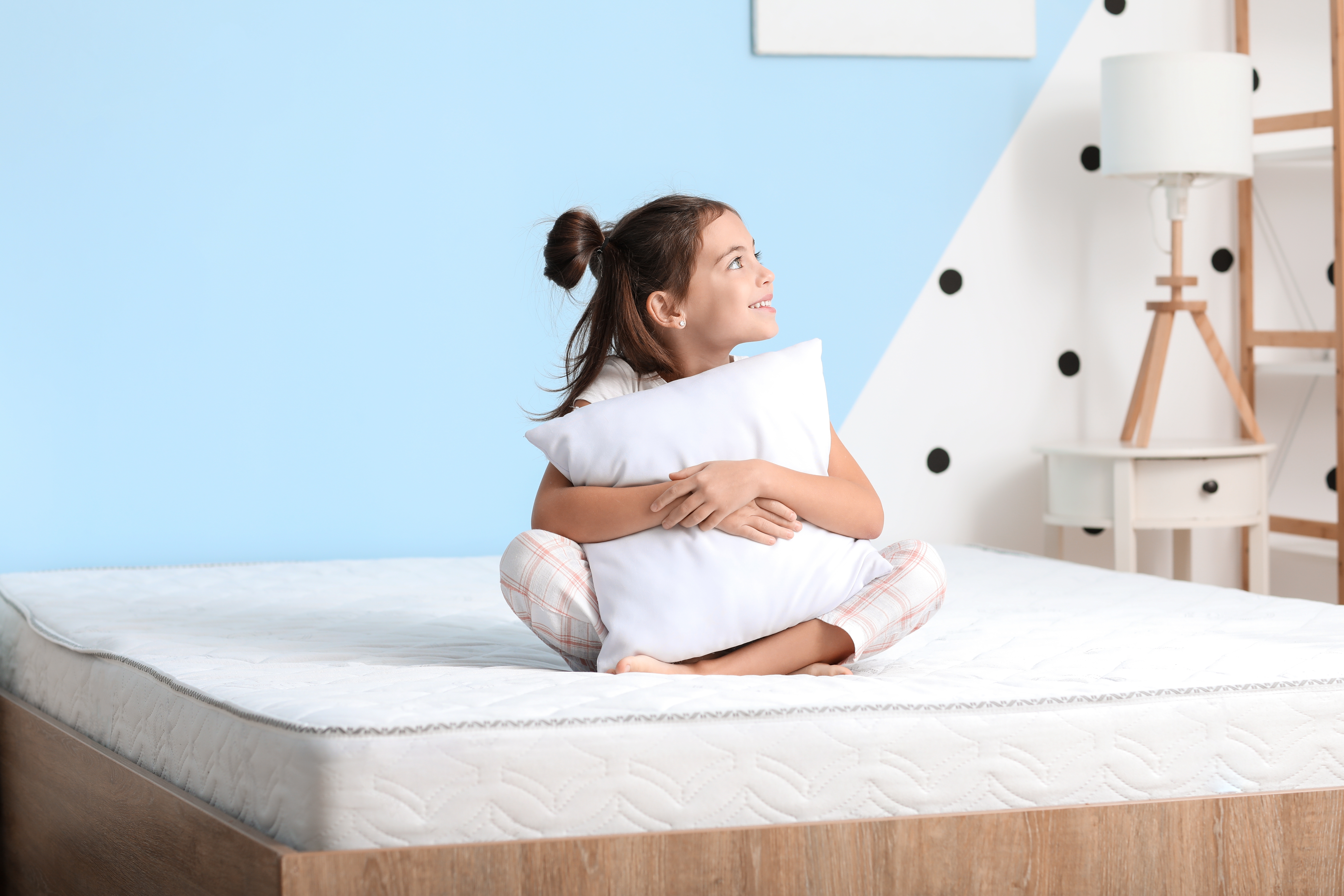 Young girl with brown hair sits on a mattress hugging a pillow