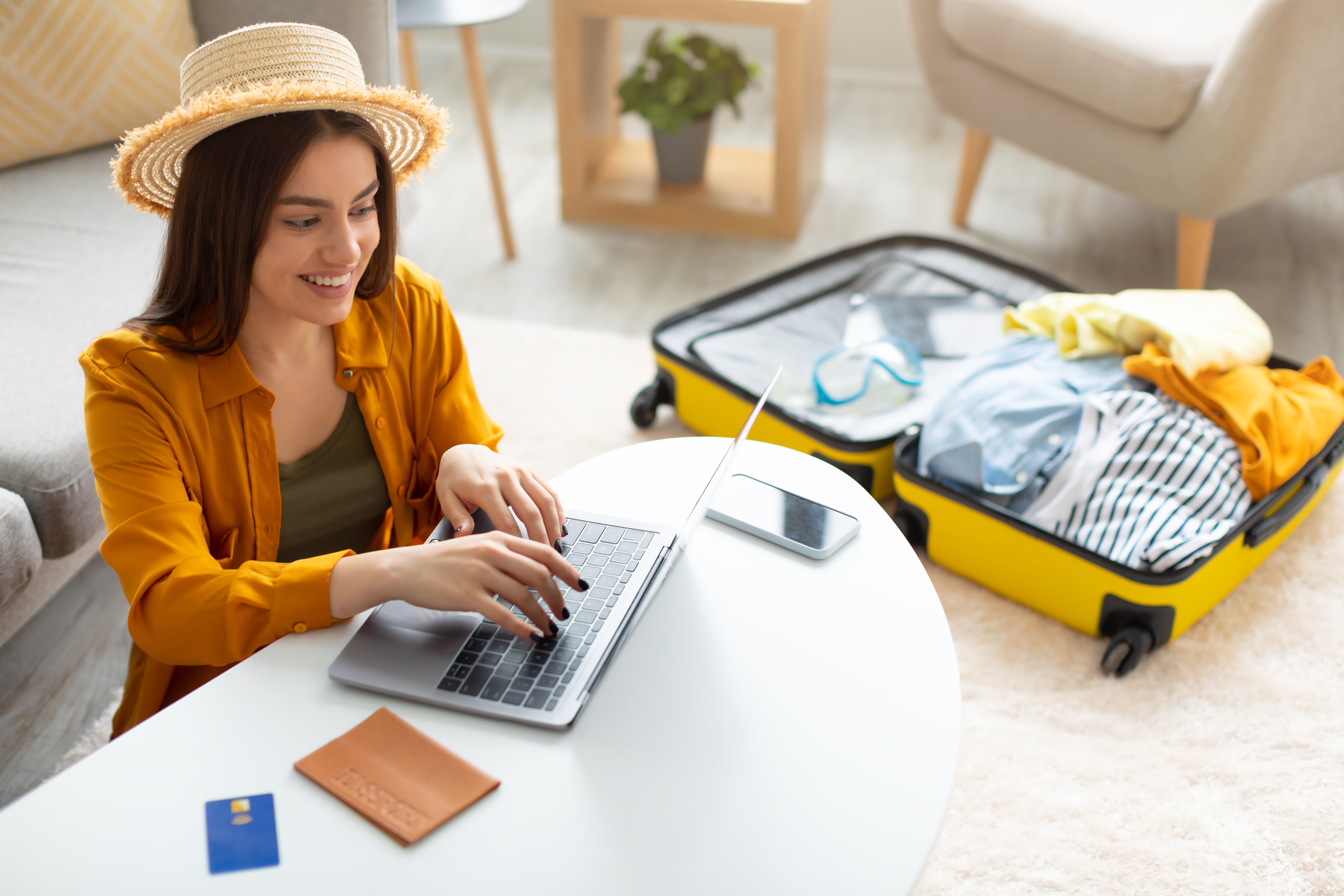 Woman wearing a hat booking a vacation on her computer next to an open suitcase full of clothes
