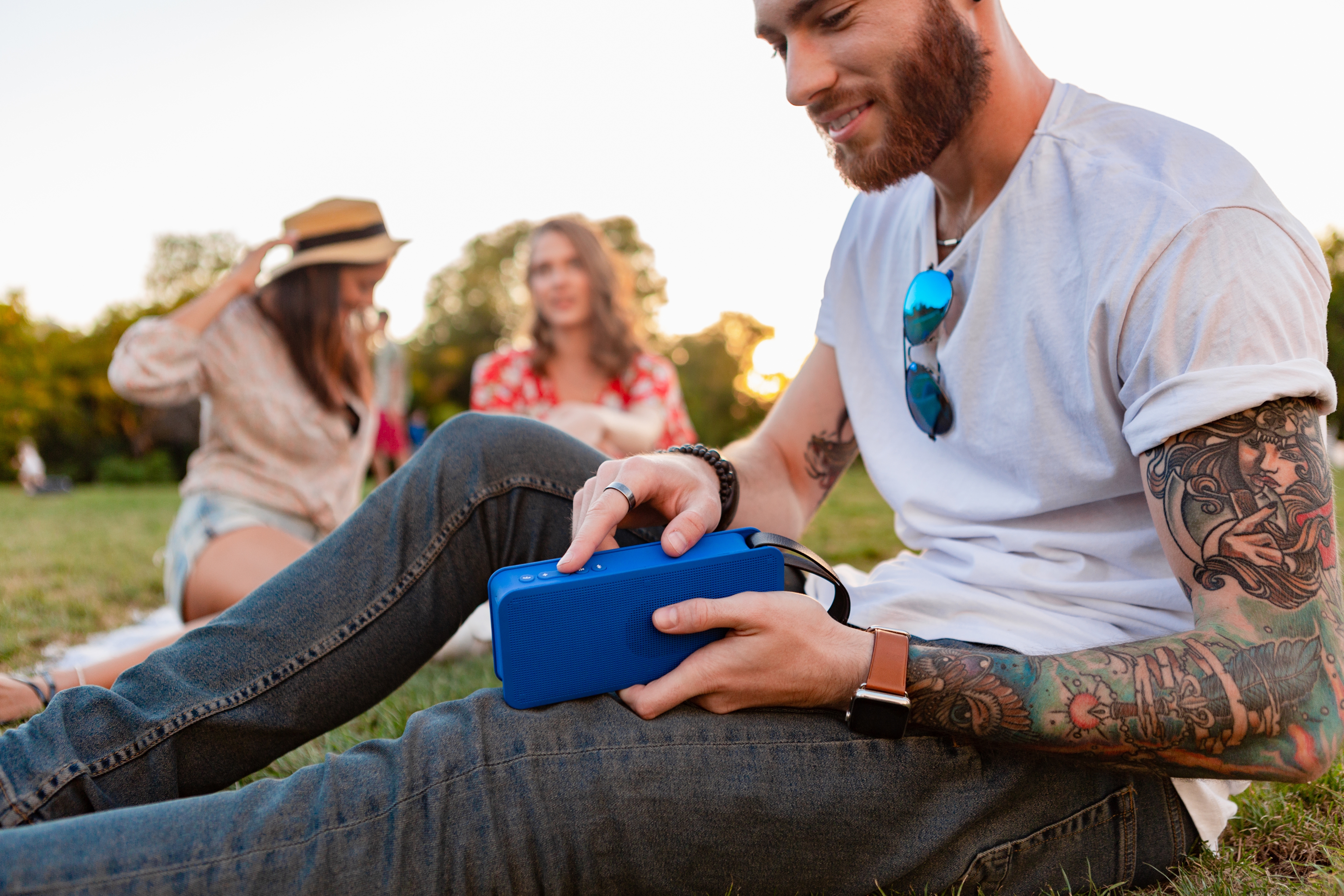 Man and two women sitting in a park listening to music on a blue outdoor speaker
