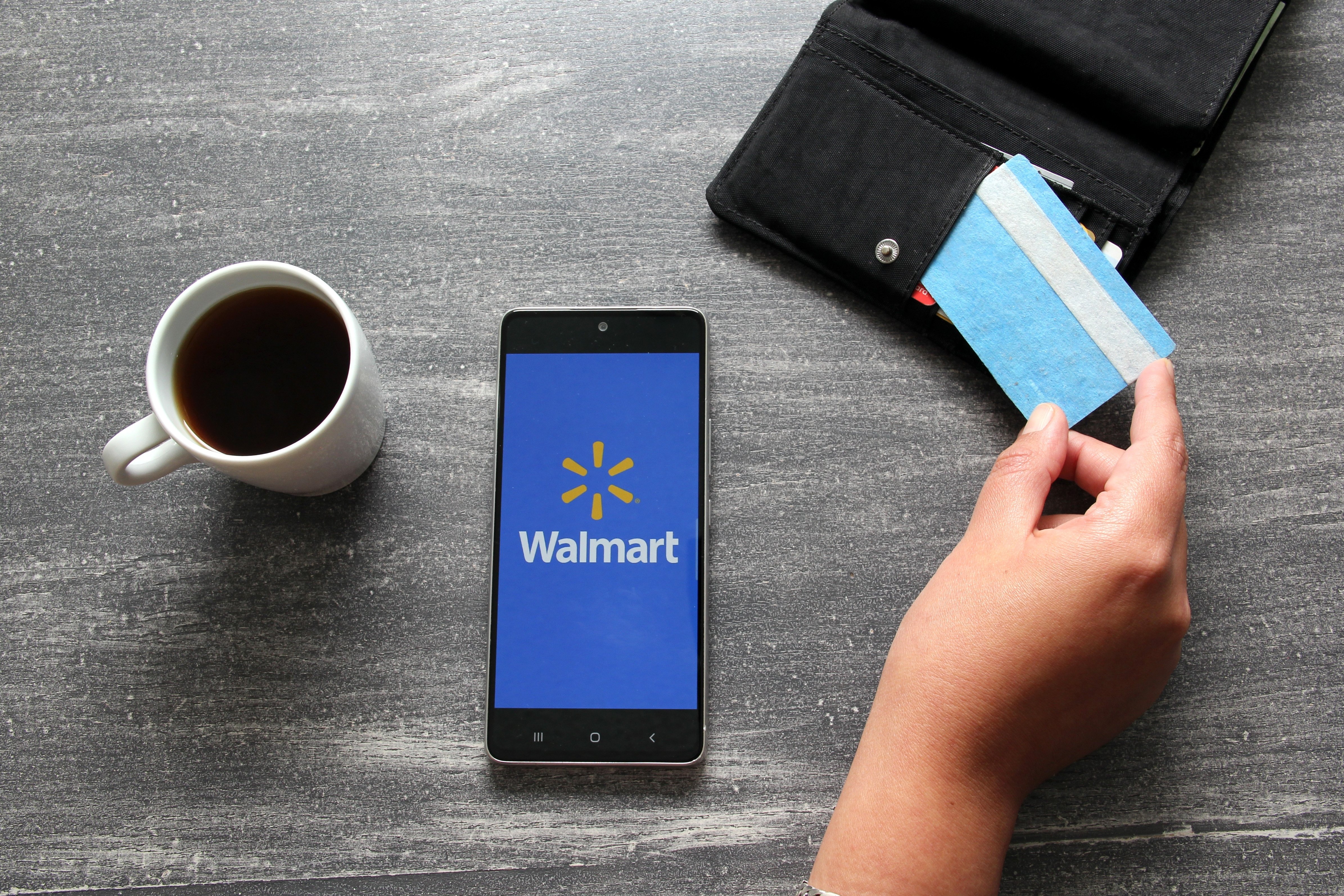 Person sitting over a cup of coffee with their smartphone showing the Walmart logo and holding their credit card