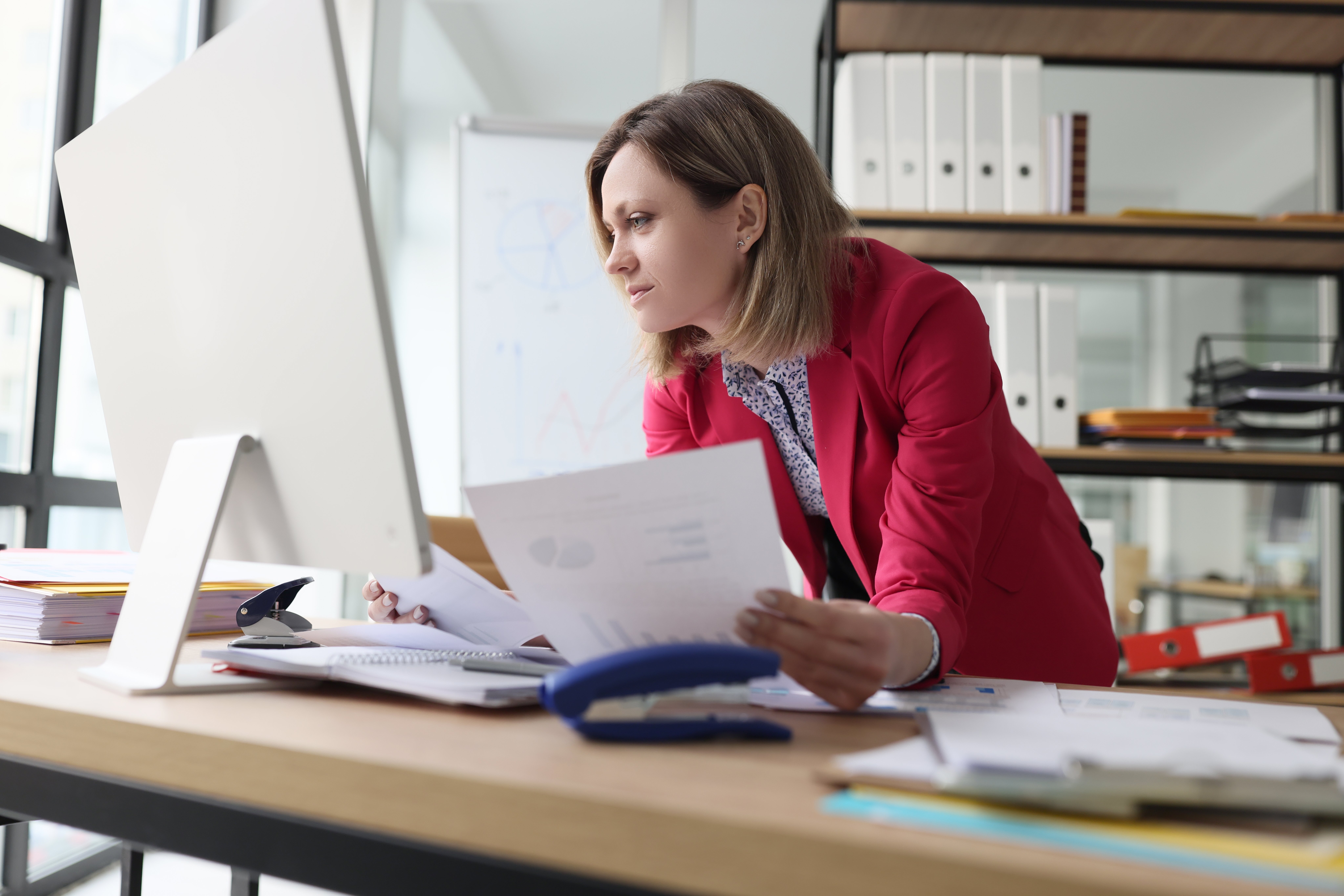 Woman wearing a red jacket leaning across a desk holding papers