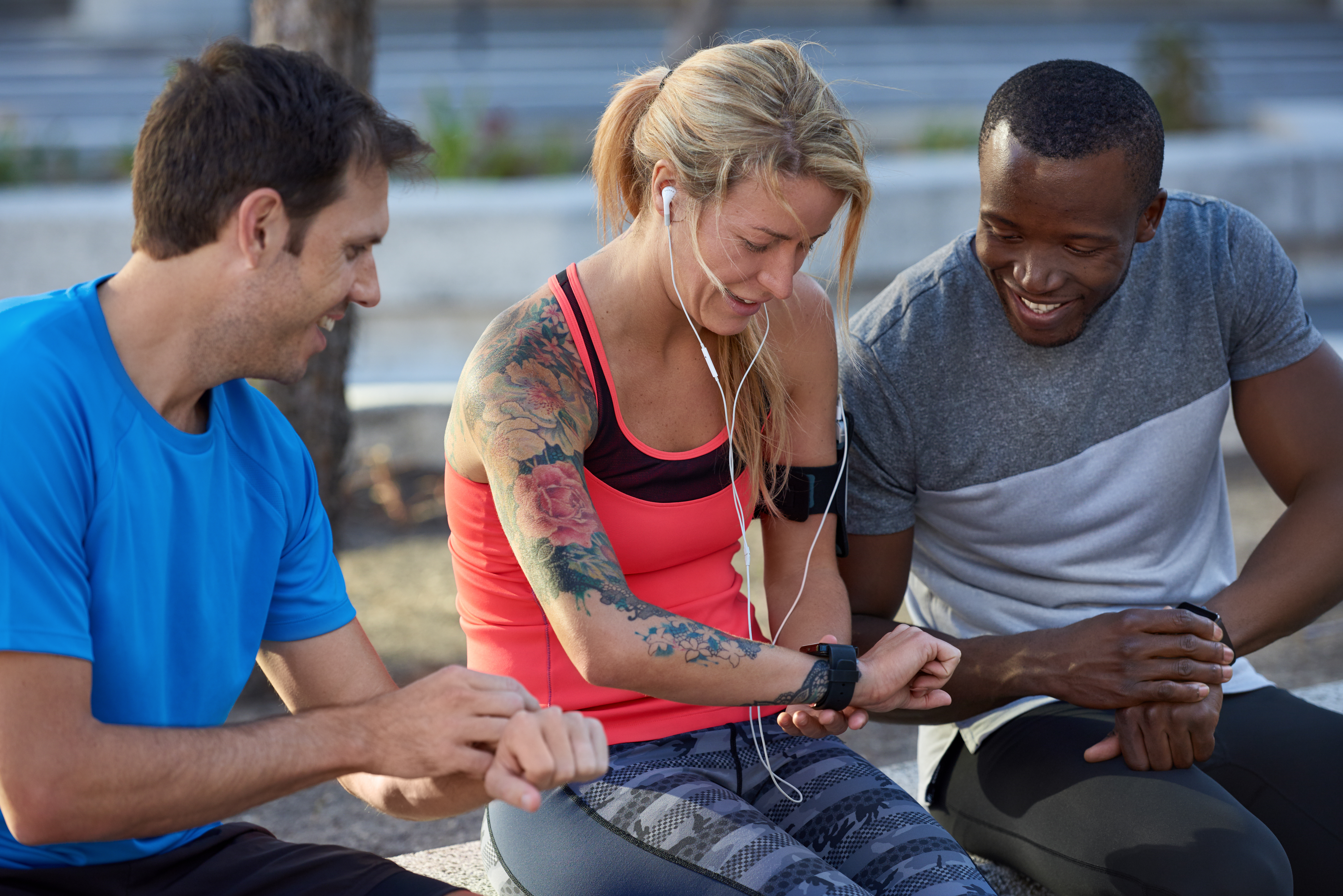 Two men and a woman sitting on a bench looking at their fitness trackers