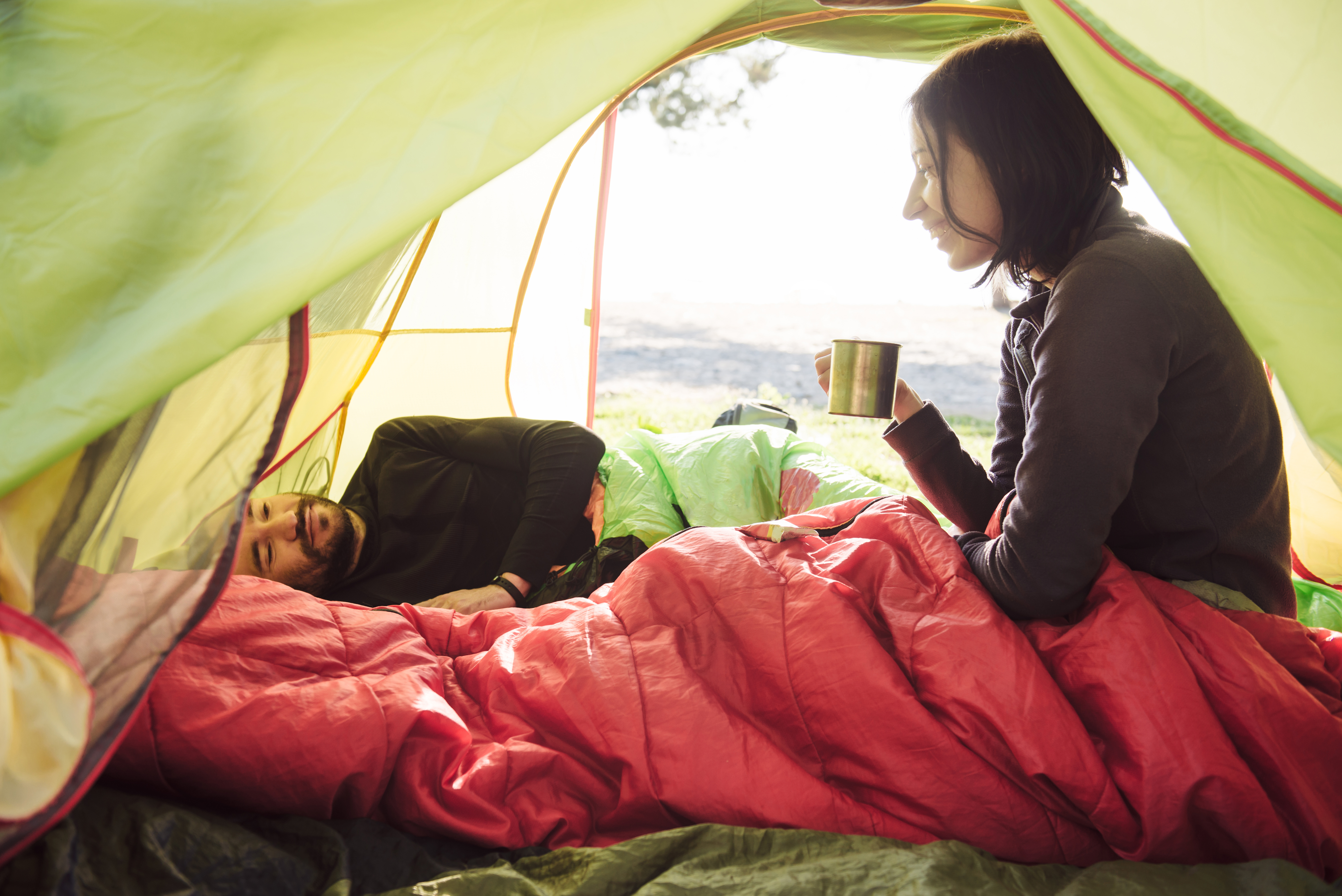 Woman in a red sleeping bag sitting up in her tent drinking from a mug